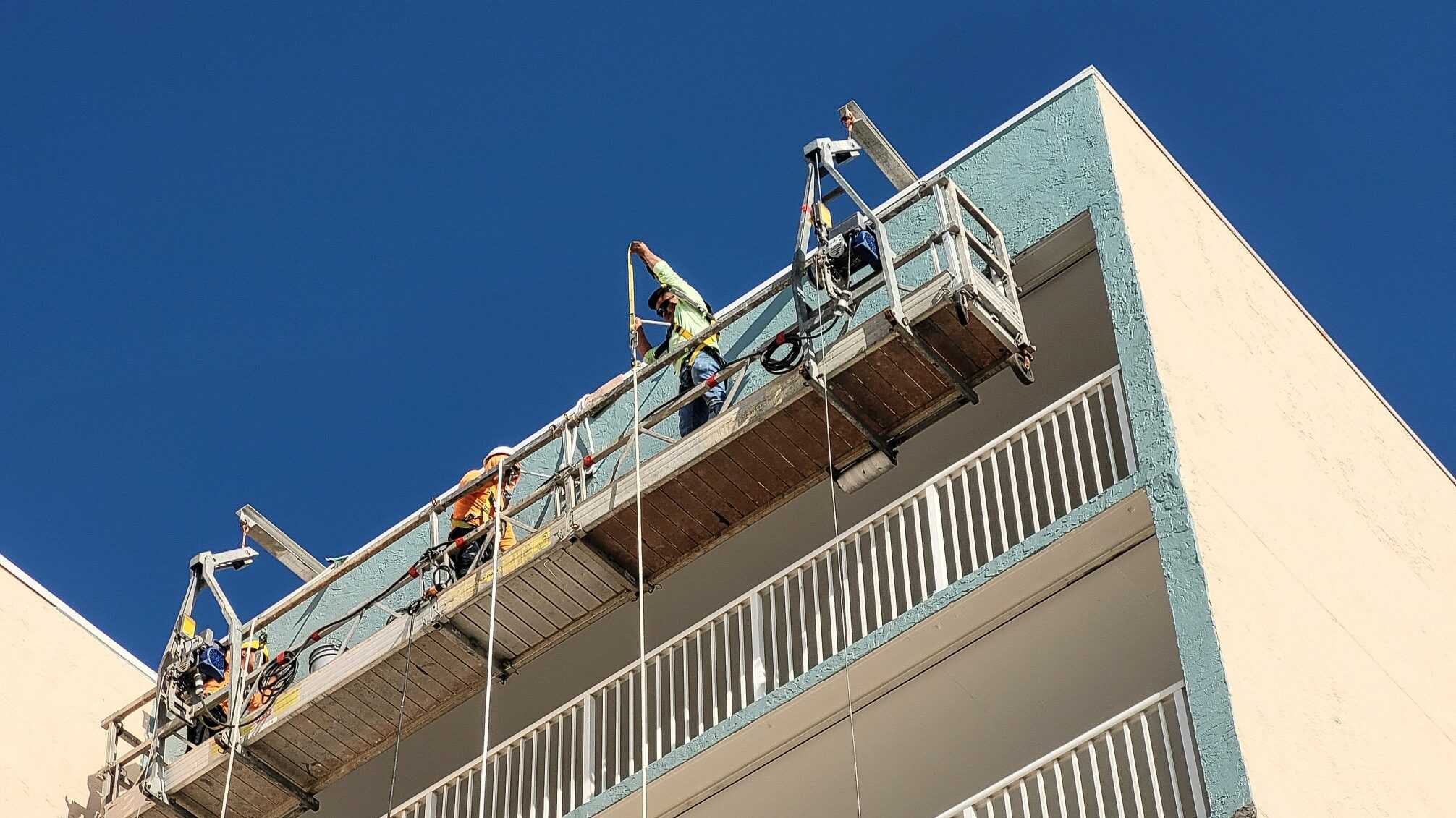 ethnic workers in hard hats on platform with ropes and pulleys painting the outside of a beach condo t20 aaZp36 edited - Fall Protection VS Fall Prevention