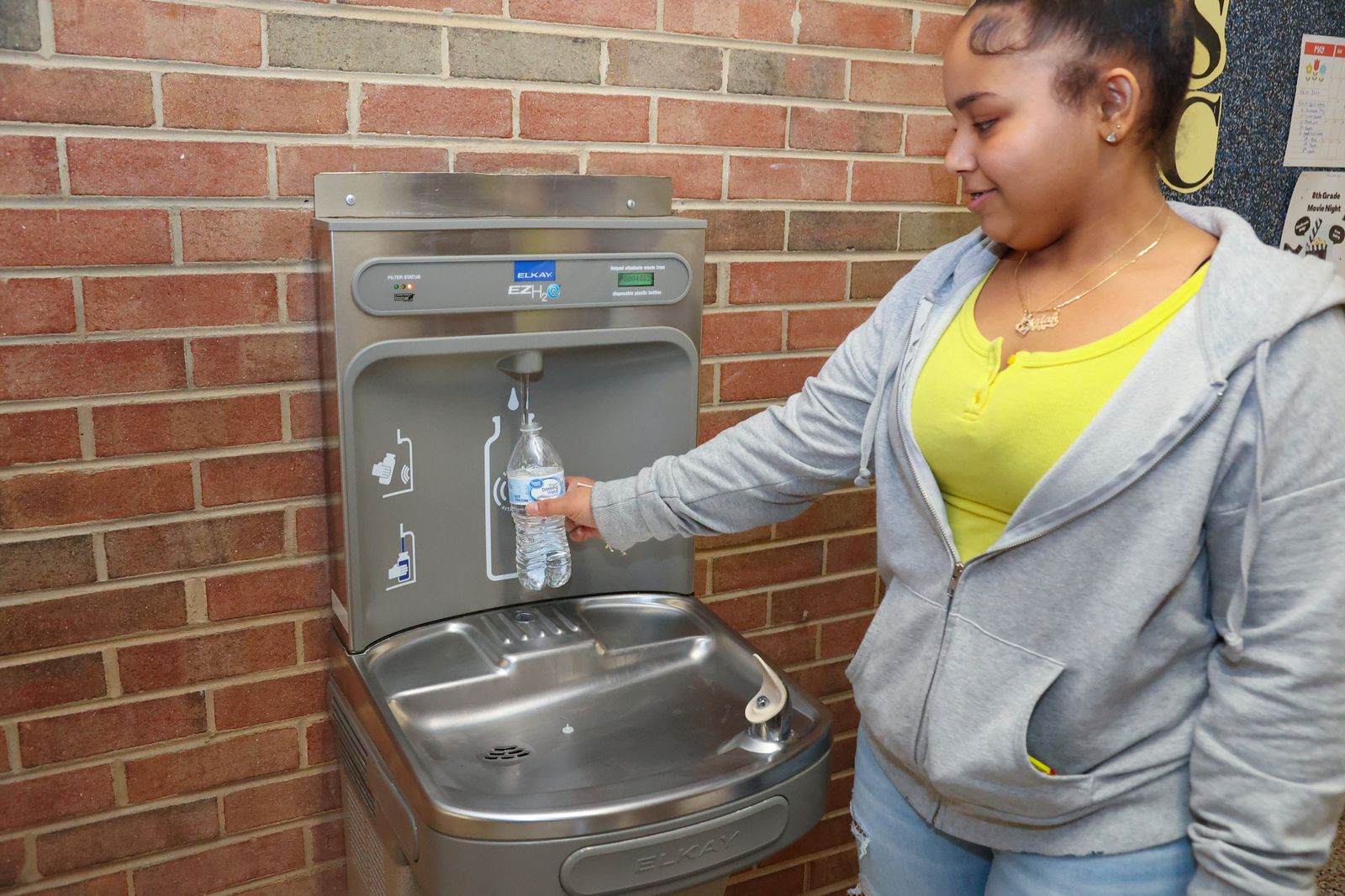 A student stands near a water fountain that has a spout for water bottles. The student is filling a bottle with water. The fountain i against a brick wall.